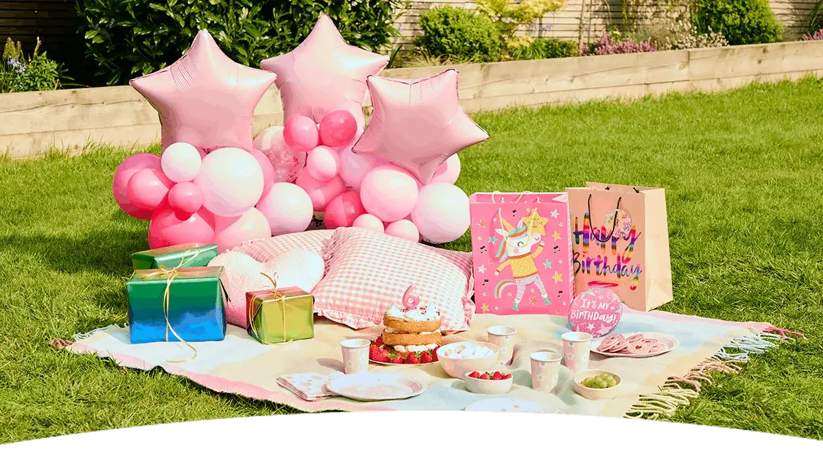 Pastel birthday picnic setup with pink balloons, gift bags, a birthday cake, party tableware, cushions, and a number 6 birthday decoration on a garden lawn. Pastel birthday picnic setup with pink balloons, gift bags, a birthday cake, party tableware, cushions, and a number 6 birthday decoration on a garden lawn.
