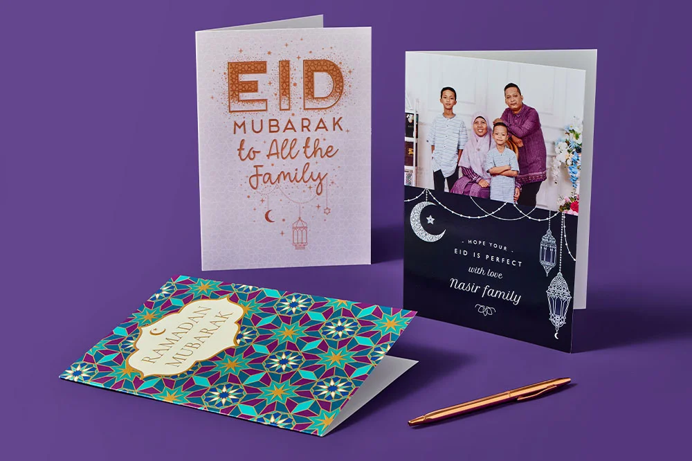 Three Ramadan and Eid greeting cards displayed on a purple background: a geometric patterned ‘Ramadan Mubarak’ card, a glitter‑style ‘Eid Mubarak to All the Family’ card, and a personalised card with lantern designs, with a pen placed beside them.
