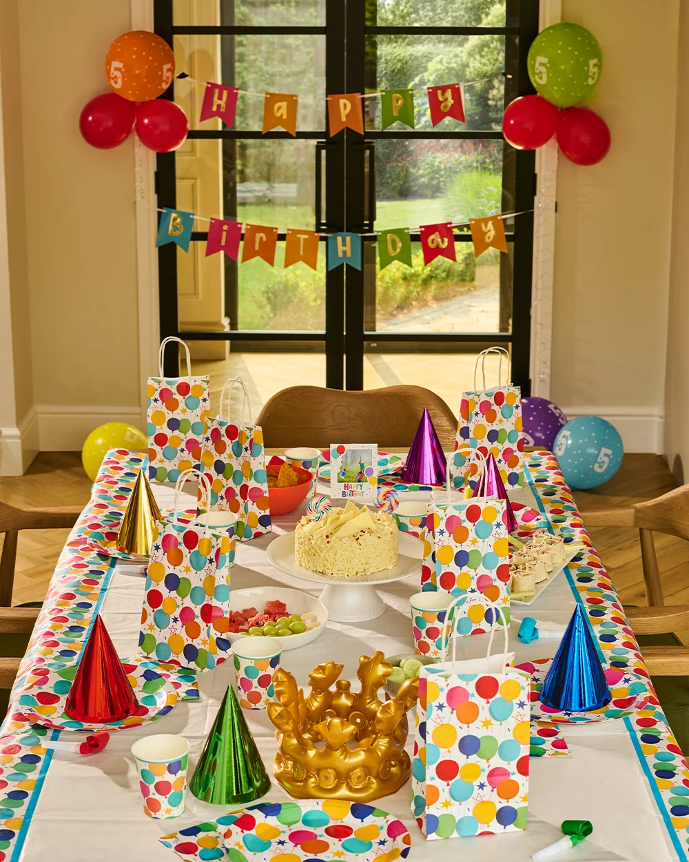 A colourful birthday party table decorated with bright balloon-print tableware, matching gift bags and multicoloured party hats. The table features a birthday cake on a stand, party food, cups and napkins, with a gold crown centrepiece in the foreground. In the background, a ‘Happy Birthday’ bunting banner hangs across glass doors, flanked by clusters of numbered balloons and additional party decorations, creating a vibrant birthday celebration setup.