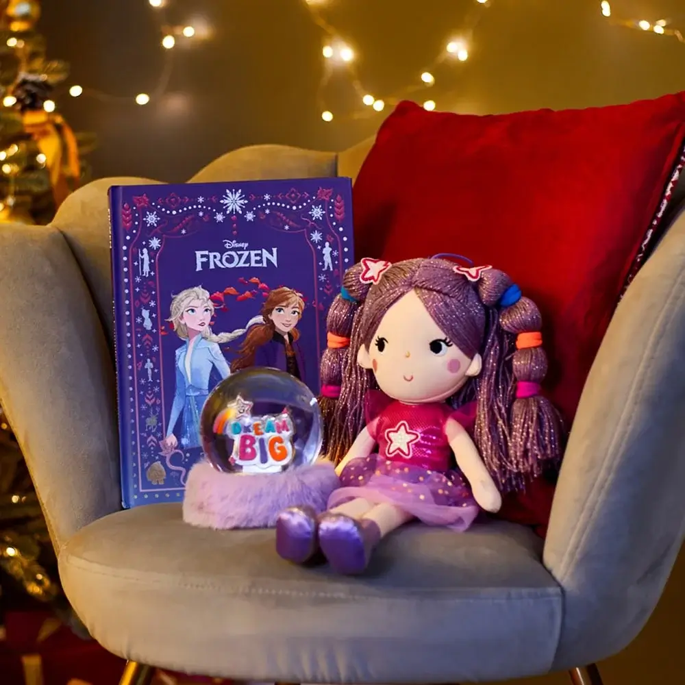 Cozy chair with a red cushion displaying a Disney Frozen book with Elsa and Anna, a plush doll with purple hair and a star on its dress, and a snow globe with Dream Big on a fluffy base, set against a background of warm fairy lights.