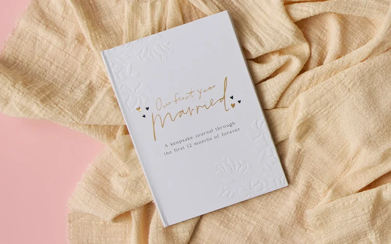 A white keepsake journal titled ‘Our First Year Married’ with gold script lettering and embossed floral details, placed on soft beige fabric against a pink background. A white keepsake journal titled ‘Our First Year Married’ with gold script lettering and embossed floral details, placed on soft beige fabric against a pink background.