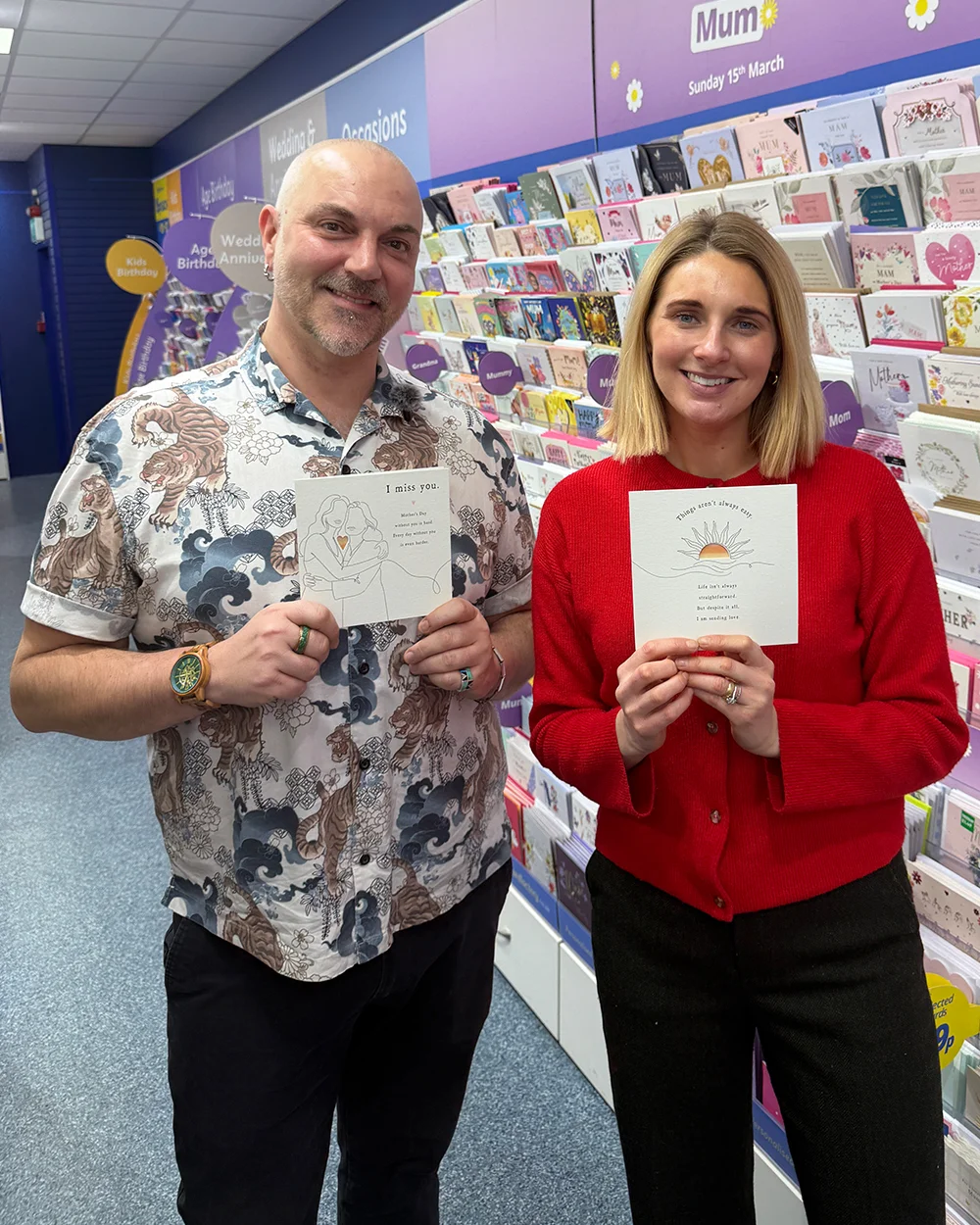 Two people standing in a greeting‑card aisle, each holding a square Mother’s Day card. One card features a line‑art drawing with a small heart cut‑out and the message ‘I miss you.’ The other shows a sun illustration with a supportive message. Rows of cards are displayed on shelves behind them.