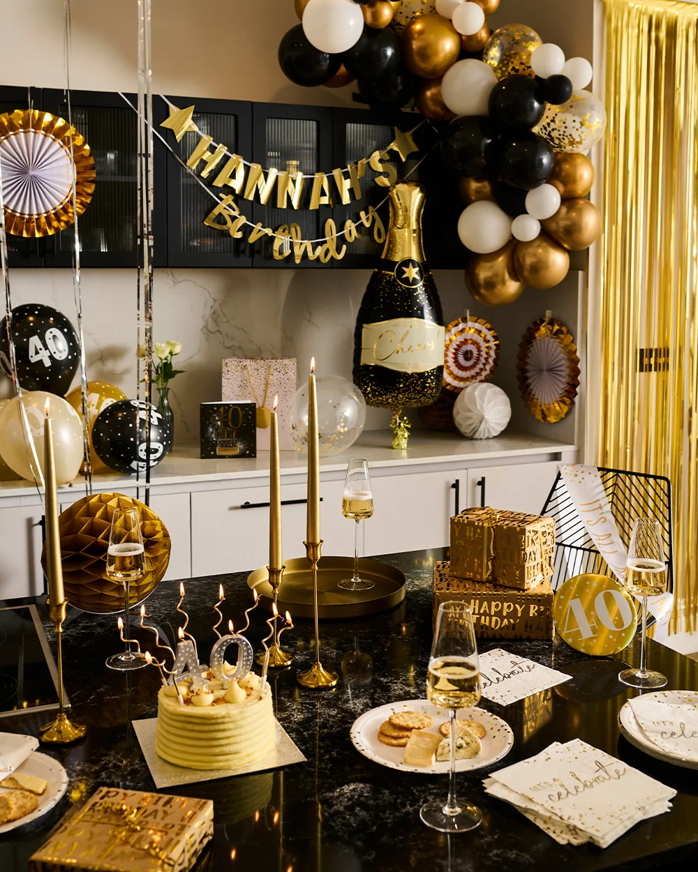 A black, gold and white milestone birthday party setup featuring a balloon garland, gold streamers and a personalised banner reading ‘Hannah’s Birthday.’ The display includes a large champagne bottle balloon, number 40 decorations, wrapped gifts, party tableware and glasses filled with sparkling drink. In the foreground, a birthday cake topped with lit number 40 candles sits on a black table surrounded by gold candle holders, plates, napkins and celebration decorations.