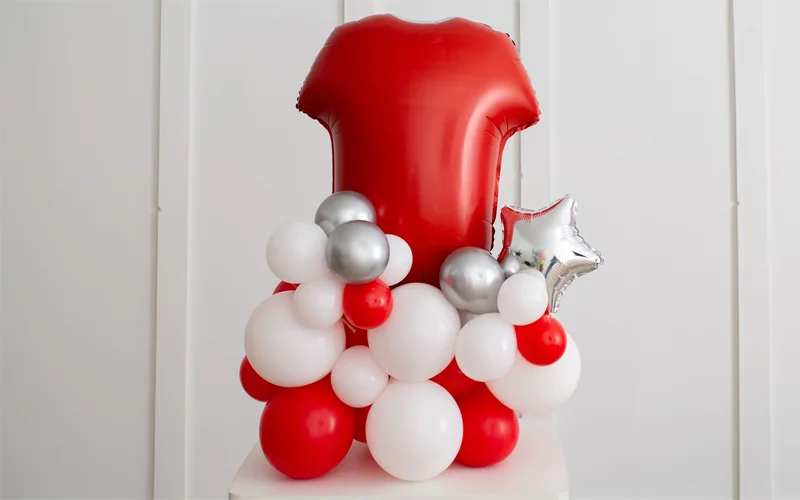A balloon centrepiece featuring a large red number 1 foil balloon attached to a base of red, white and silver balloons, accented with a silver star balloon, displayed on a white plinth against a white panelled wall. A balloon centrepiece featuring a large red number 1 foil balloon attached to a base of red, white and silver balloons, accented with a silver star balloon, displayed on a white plinth against a white panelled wall.