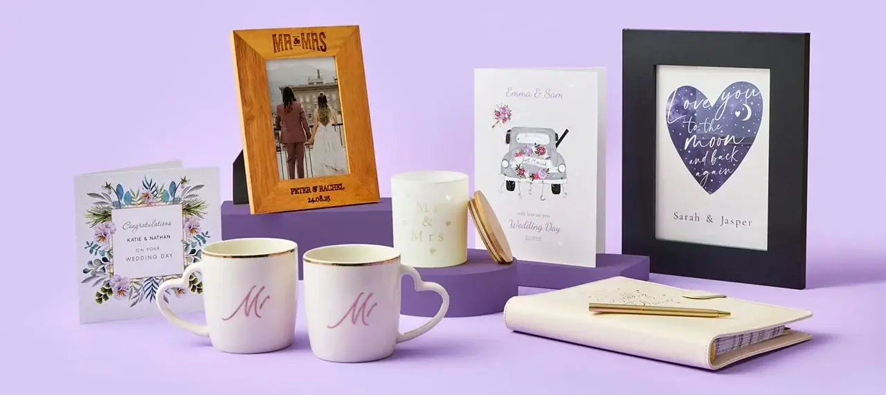 A collection of wedding-themed items displayed on a purple surface. The items include two greeting cards, two mugs with the letter 'M' printed on them, a framed photo of a couple labeled 'MR & MRS', a candle with the text 'Mr & Mrs', and another framed print with the text 'Love you to the moon and back'. There is also an open notebook with a gold pen resting on it.