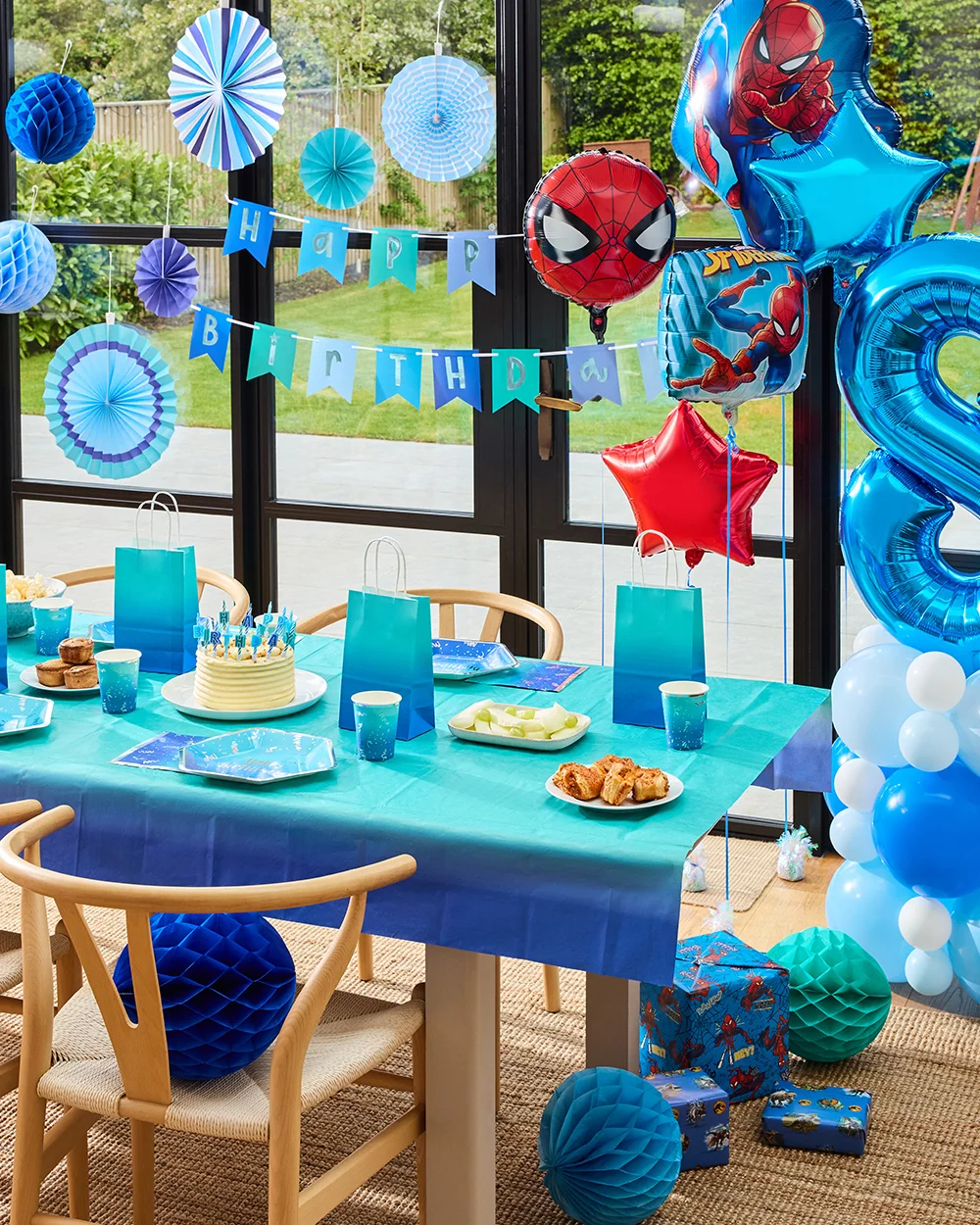 A Spider-Man themed birthday party table decorated in shades of blue, featuring matching plates, cups, napkins and gift bags arranged around a birthday cake. Blue paper fan decorations and a ‘Happy Birthday’ banner hang in the background, while Spider-Man foil balloons and a large blue number 8 balloon display complete the celebration setup.