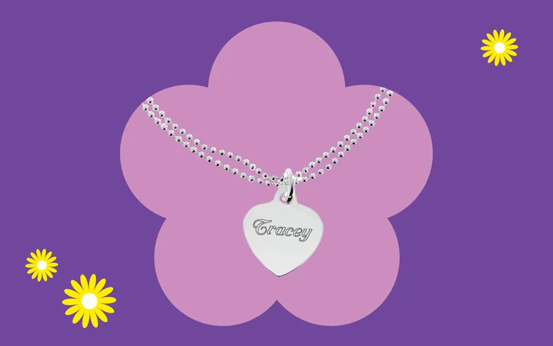 A silver heart‑shaped pendant engraved with the name ‘Tracey,’ displayed on a beaded chain against a purple background with a pink flower shape.