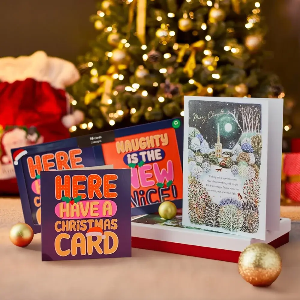Festive scene with a decorated Christmas tree and three Christmas cards featuring bold text, playful messages, and a snowy village illustration. Festive scene with a decorated Christmas tree and three Christmas cards featuring bold text, playful messages, and a snowy village illustration.