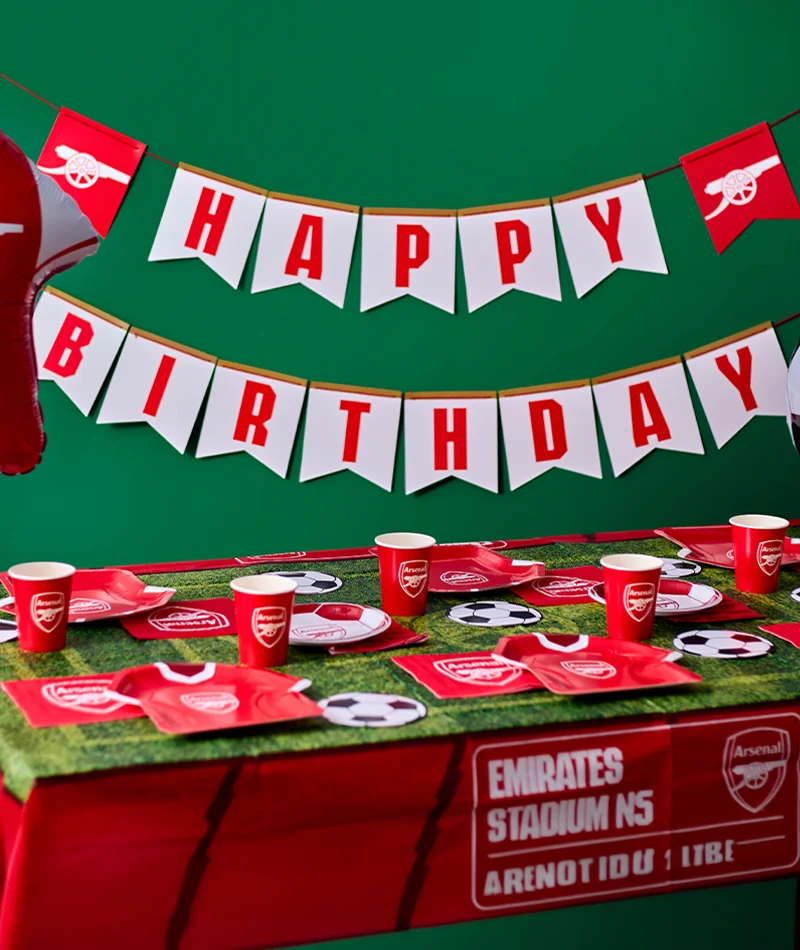 An Arsenal football-themed birthday party table display featuring red branded plates, cups and napkins arranged on a grass-effect table covering. A ‘Happy Birthday’ bunting banner hangs against a green background, with football-shaped balloons at either side and football-themed table decorations completing the celebration setup. An Arsenal football-themed birthday party table display featuring red branded plates, cups and napkins arranged on a grass-effect table covering. A ‘Happy Birthday’ bunting banner hangs against a green background, with football-shaped balloons at either side and football-themed table decorations completing the celebration setup.