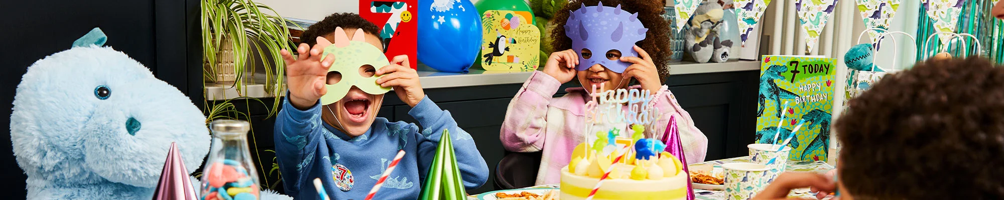 A dinosaur-themed birthday party table with themed cups, plates and decorations, featuring a birthday cake topped with a ‘Happy Birthday’ sign and a number 7 candle. Dinosaur masks, party hats, dinosaur bunting, balloons and a personalised birthday card are displayed around the table as children take part in the celebration.
