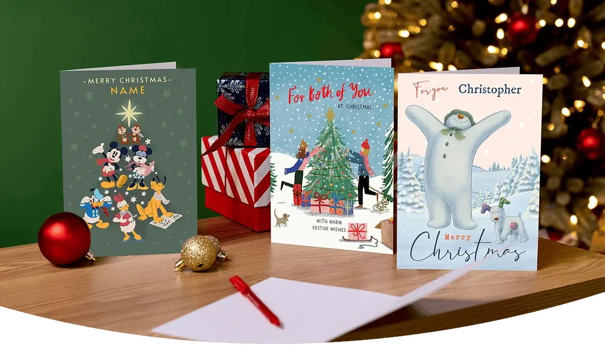 Selection of Christmas cards, including Disney, festive and personalised designs, displayed on a wooden table with wrapped presents and a decorated Christmas tree in the background.