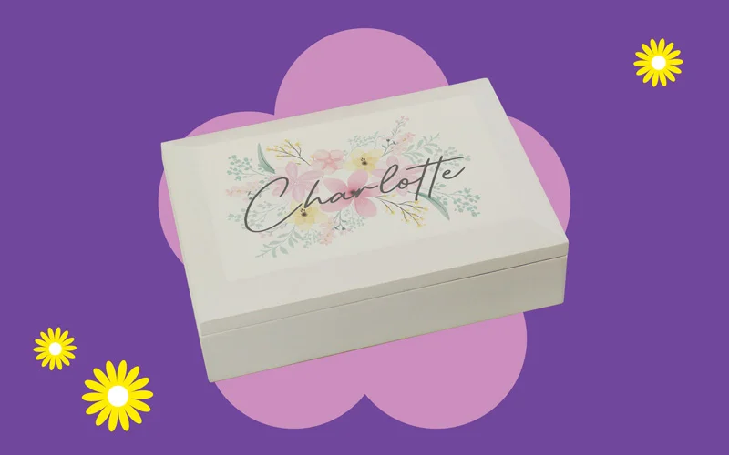 A white keepsake box with pastel floral artwork and the name ‘Charlotte’ on the lid, shown against a purple background with yellow flower graphics.