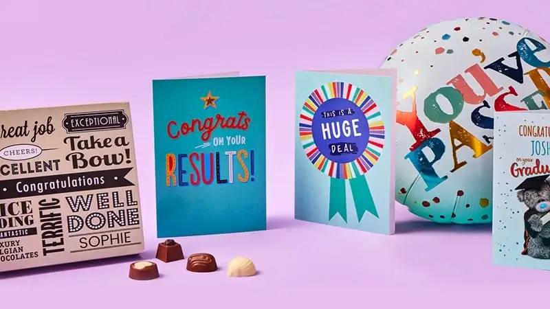 Celebratory arrangement featuring congratulatory cards, a box of chocolates with phrases like 'Great job' and 'Well Done', a graduation card for Joshua, and a balloon that says 'You've PASSED'