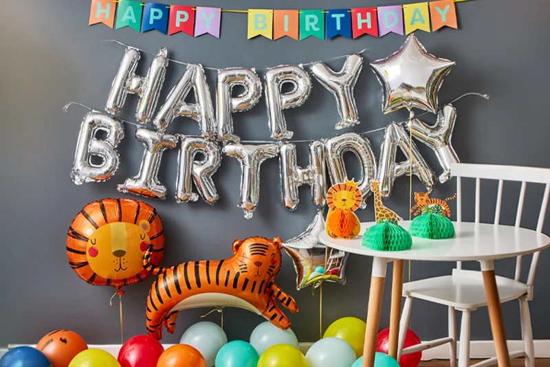 A jungle-themed birthday party display featuring silver foil ‘Happy Birthday’ balloons, a colourful birthday bunting banner, animal-shaped foil balloons including a lion and tiger, and a small party table decorated with safari animal centrepieces and honeycomb decorations. Multicoloured balloons are scattered across the floor to complete the celebration setup.