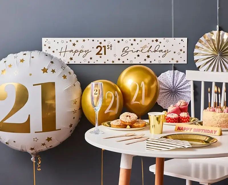 21st birthday celebration setup with gold and white balloons, a champagne flute, a banner, and a table decorated with cake, cupcakes, donuts, and party items. 21st birthday celebration setup with gold and white balloons, a champagne flute, a banner, and a table decorated with cake, cupcakes, donuts, and party items.