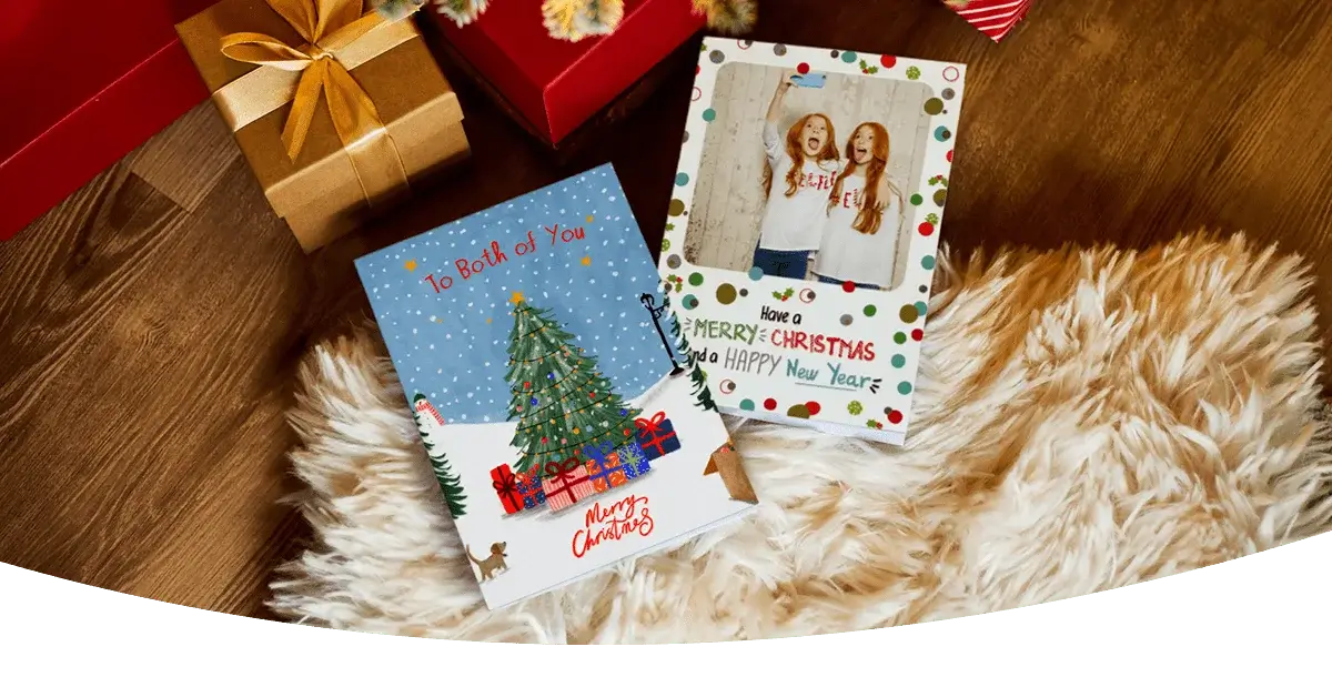 Two Christmas cards placed on a fluffy rug next to wrapped gifts. One card features a decorated Christmas tree with presents and the text To Both of You Merry Christmas, while the other has a festive design with colorful dots and the message Have a Merry Christmas and a Happy New Year.