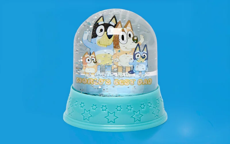 A snow globe featuring Bluey family characters inside with confetti pieces, mounted on a teal base decorated with star shapes, with the text ‘World’s Best Dad’ printed on the globe, displayed against a blue background. A snow globe featuring Bluey family characters inside with confetti pieces, mounted on a teal base decorated with star shapes, with the text ‘World’s Best Dad’ printed on the globe, displayed against a blue background.