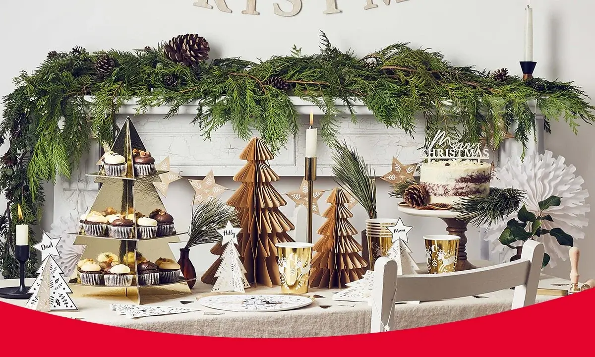 Festive Christmas table setting with paper trees, candles, greenery, cupcakes, a cake with Merry Christmas topper, personalized tree-shaped place cards, and a decorated mantel with pinecones and evergreen branches. Festive Christmas table setting with paper trees, candles, greenery, cupcakes, a cake with Merry Christmas topper, personalized tree-shaped place cards, and a decorated mantel with pinecones and evergreen branches.