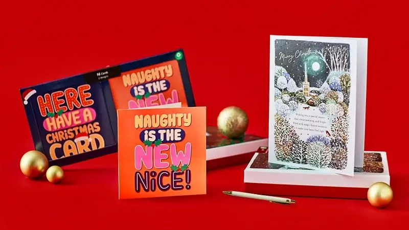 A festive display of three Christmas cards on a red background, featuring bold text designs and a winter scene illustration, surrounded by gold ornaments and a pen