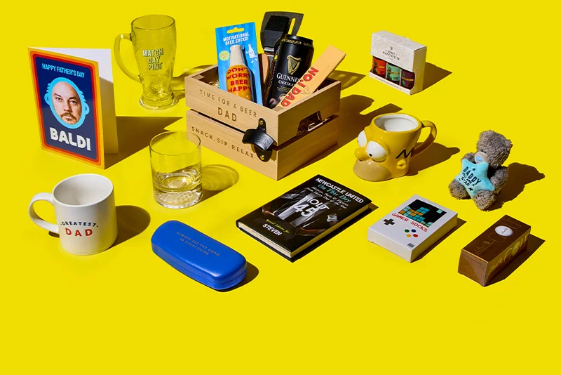 A bright yellow flat‑lay display of Father’s Day gifts, including a wooden crate engraved ‘Time for a Beer Dad – Snack, Sip, Relax,’ filled with items such as a drink can, chocolate bar and novelty tools, surrounded by a variety of gifts including a ‘Happy Father’s Day’ card, a ‘Match Day Pint’ glass, mugs, a game‑controller tin, books, sweets, and a small teddy bear wearing a ‘Best Dad’ outfit.