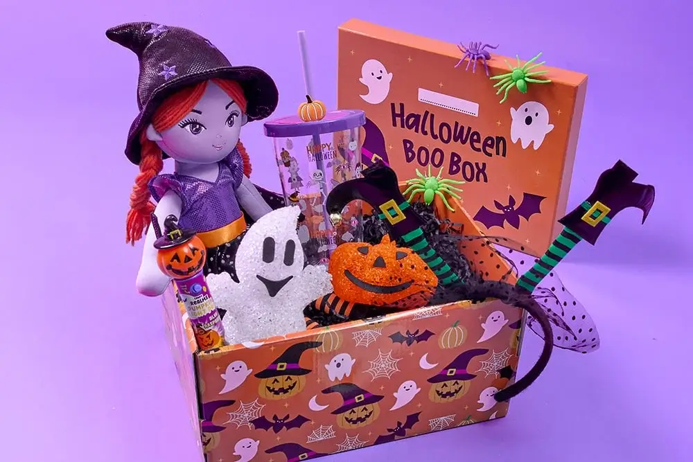 Halloween-themed gift box with witch doll, ghost and pumpkin decorations, tumbler cup, and festive accessories on a purple background.