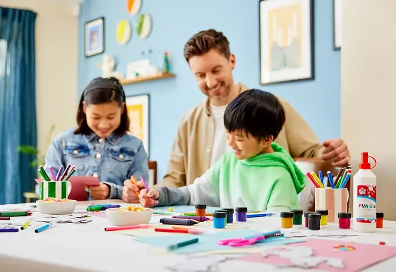 An adult and two children sitting at a table doing arts and crafts with paints, paper and pens in a bright room.