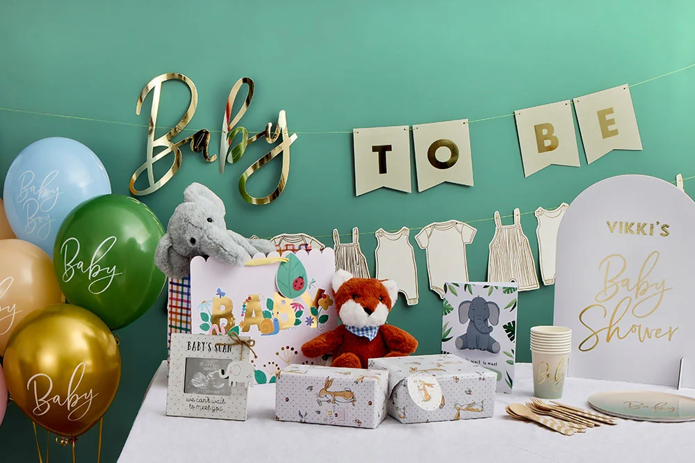 A baby shower table decorated with pastel balloons, a gold ‘Baby’ sign, ‘TO BE’ bunting and baby clothes garland. The display includes wrapped gifts, soft toy animals, personalised tableware, baby shower signage reading ‘Vikki’s Baby Shower,’ and coordinating baby-themed decorations in green, gold, blue and pink tones.