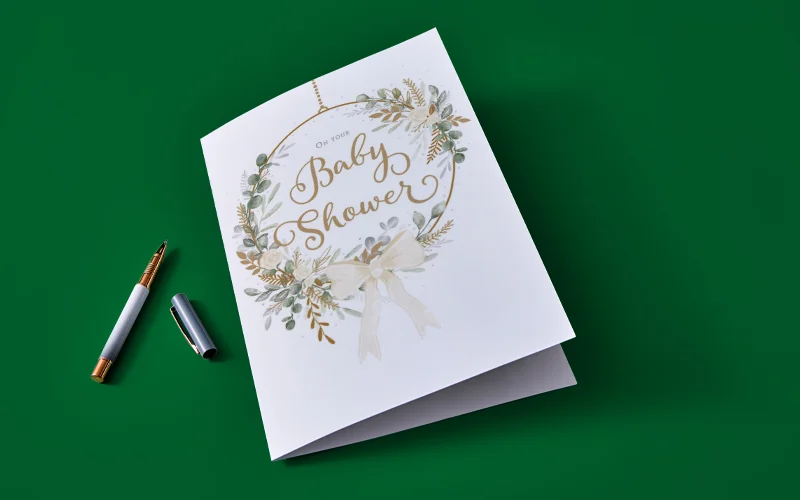 A collection of baby shower game cards displayed on a neutral background, including ‘Celebrity Baby Names’, ‘Baby Predictions’, ‘Name the Baby’ and ‘Baby Trivia’, alongside an answer sheet, pen and decorative flowers.