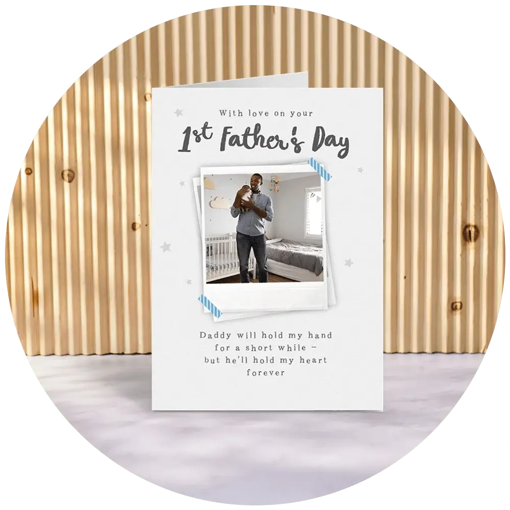A white first Father’s Day card featuring a Polaroid‑style photo frame design, small grey stars, and the text ‘With love on your 1st Father’s Day,’ with a sentimental message printed below, displayed against a wooden slatted backdrop. A white first Father’s Day card featuring a Polaroid‑style photo frame design, small grey stars, and the text ‘With love on your 1st Father’s Day,’ with a sentimental message printed below, displayed against a wooden slatted backdrop.