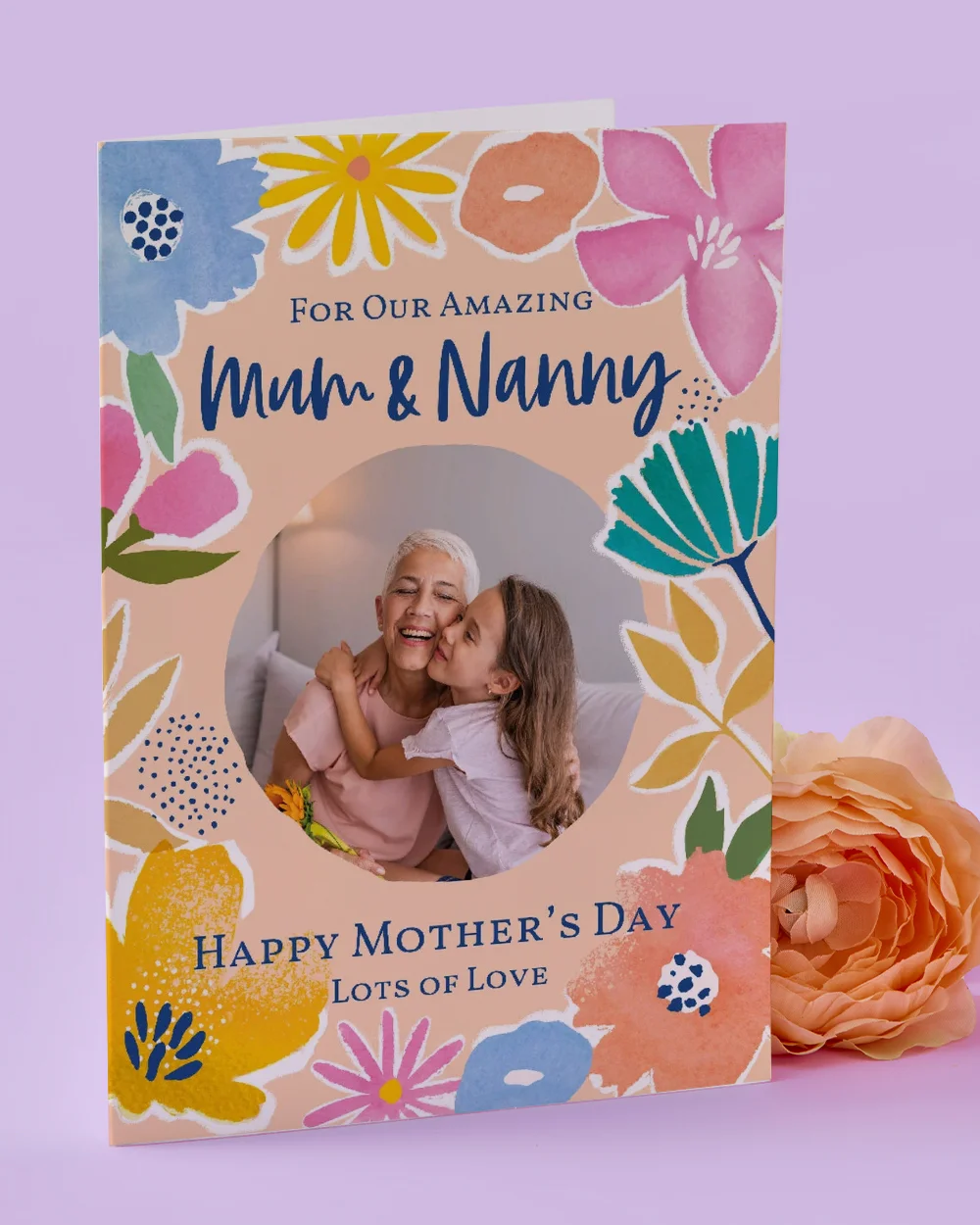 A floral Mother’s Day card titled ‘For Our Amazing Mum & Nanny,’ featuring colourful illustrated flowers and a circular photo window, displayed on a pastel purple background beside a peach flower.
