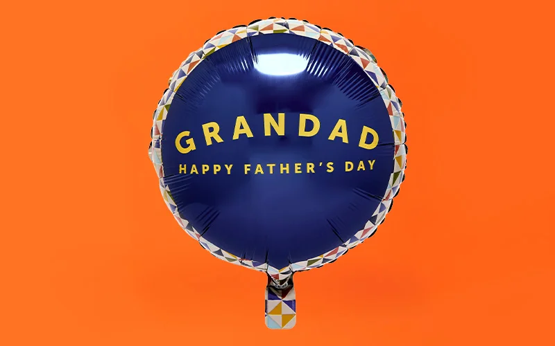 A round navy blue foil balloon with a patterned border and gold lettering reading ‘Grandad Happy Father’s Day,’ displayed against a bright orange background. A round navy blue foil balloon with a patterned border and gold lettering reading ‘Grandad Happy Father’s Day,’ displayed against a bright orange background.