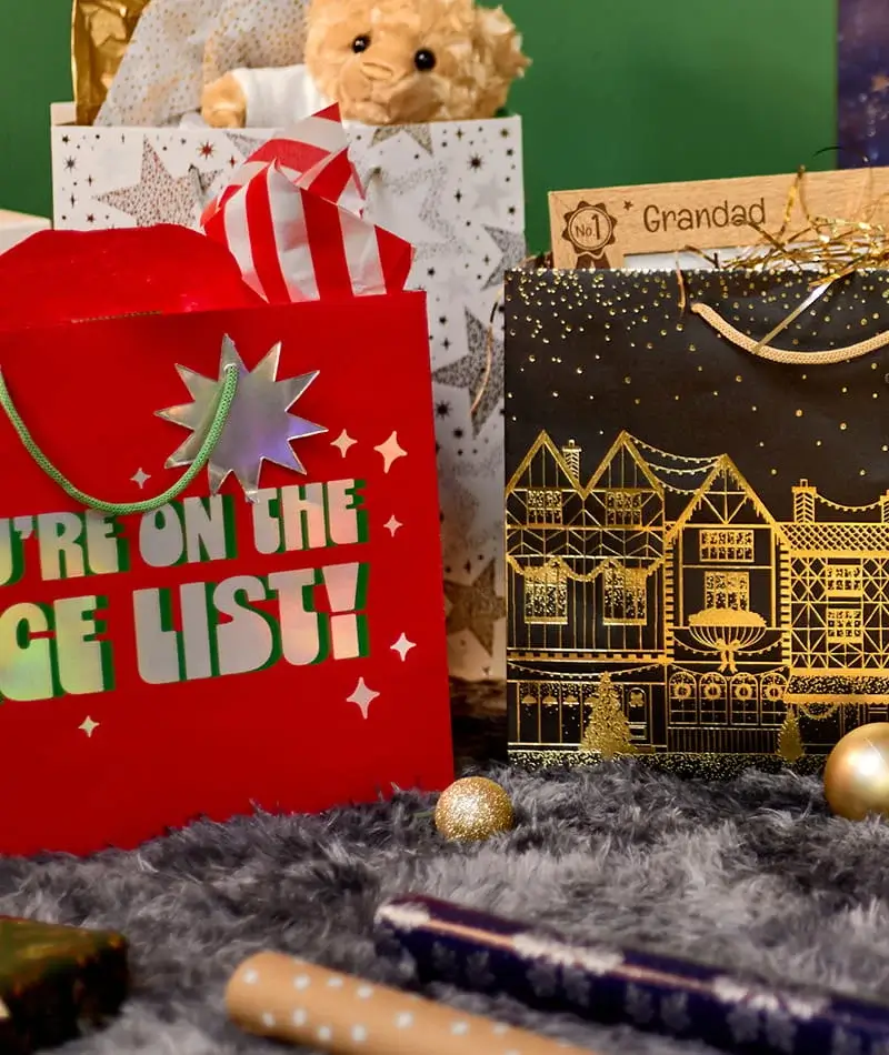 Festive Christmas gift bags filled with gifts, cards and a teddy bear, displayed on a grey rug with gold baubles and wrapped presents.