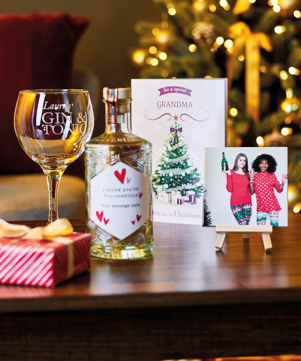 Festive Christmas scene with a personalized gin glass labeled 'Laura's GIN & TONIC', a custom gin bottle, a 'GRANDMA' Christmas card, a framed photo, and a striped gift box beside a lit tree with golden ribbons.