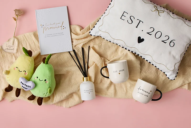 A wedding‑themed flat lay arranged on beige fabric over a pink background, featuring an ‘Our First Year Married’ journal, two pear plush toys holding a heart labelled ‘Perfect Pear,’ a reed diffuser with black sticks, two ceramic mugs printed ‘EST. 2026’ and ‘Forever starts now,’ and a decorative cushion with ‘EST. 2026’ and a small heart design.