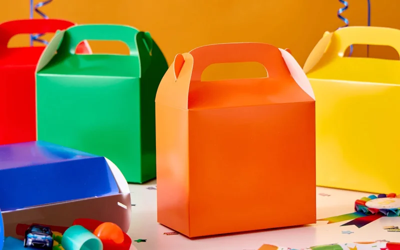 A set of colourful party favour boxes in orange, green, yellow and red, displayed on a party table with confetti and small party bag fillers against a bright orange background.