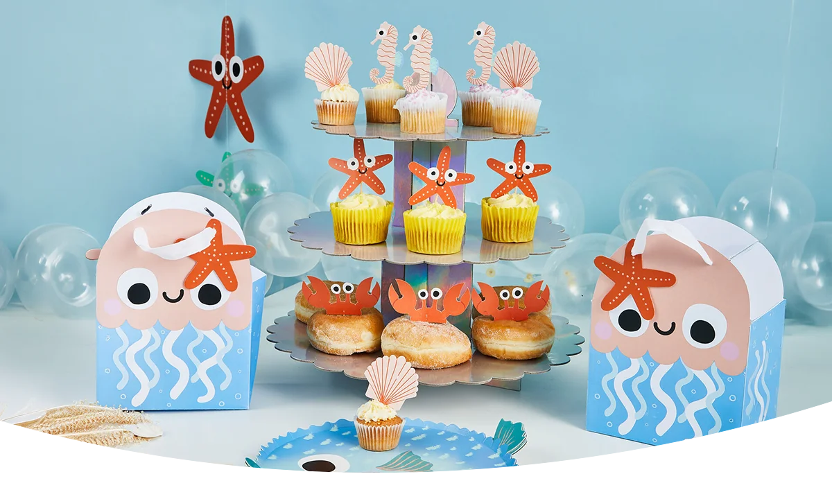 An under‑the‑sea themed party display on a light blue background, featuring a three‑tier cupcake stand with decorated cupcakes, crab‑topped donuts, jellyfish‑shaped party boxes, starfish decorations, shells, and transparent balloons arranged around the table.
