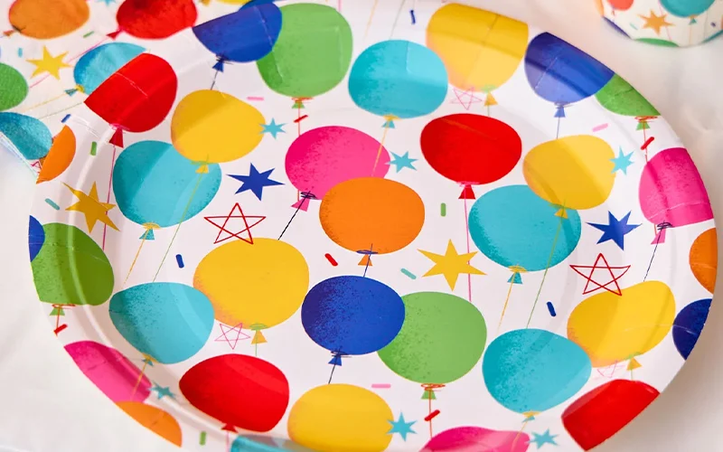 A hexagonal white party plate decorated with colourful balloon illustrations in blue, green, yellow, orange, pink and red, surrounded by star shapes and confetti details. The plate is displayed on a white table covering as part of a bright birthday party tableware collection. A hexagonal white party plate decorated with colourful balloon illustrations in blue, green, yellow, orange, pink and red, surrounded by star shapes and confetti details. The plate is displayed on a white table covering as part of a bright birthday party tableware collection.