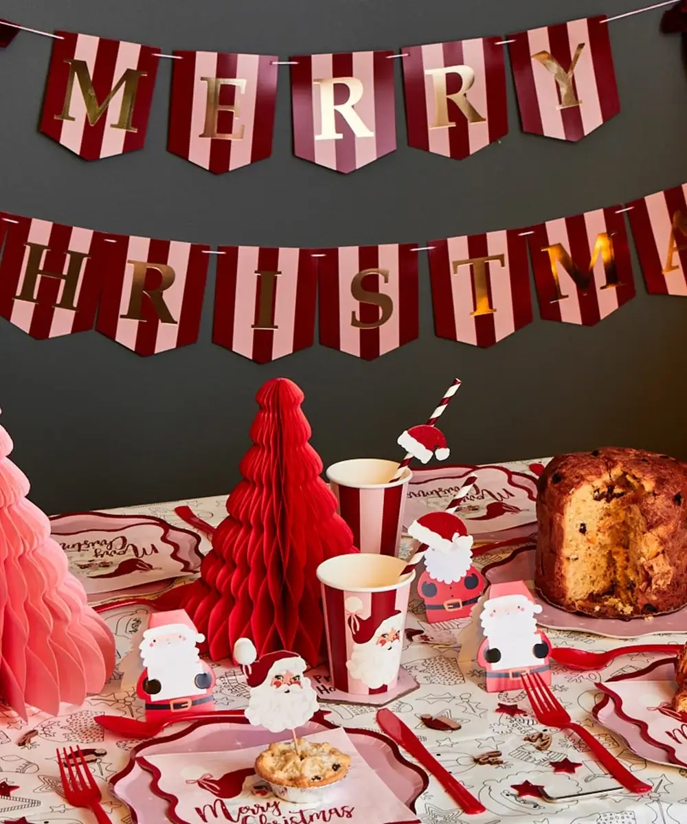 Festive table decorated with red and pink paper Christmas trees, Santa-themed cups and plates, and a large cake, with a red and gold Merry Christmas banner hanging above.