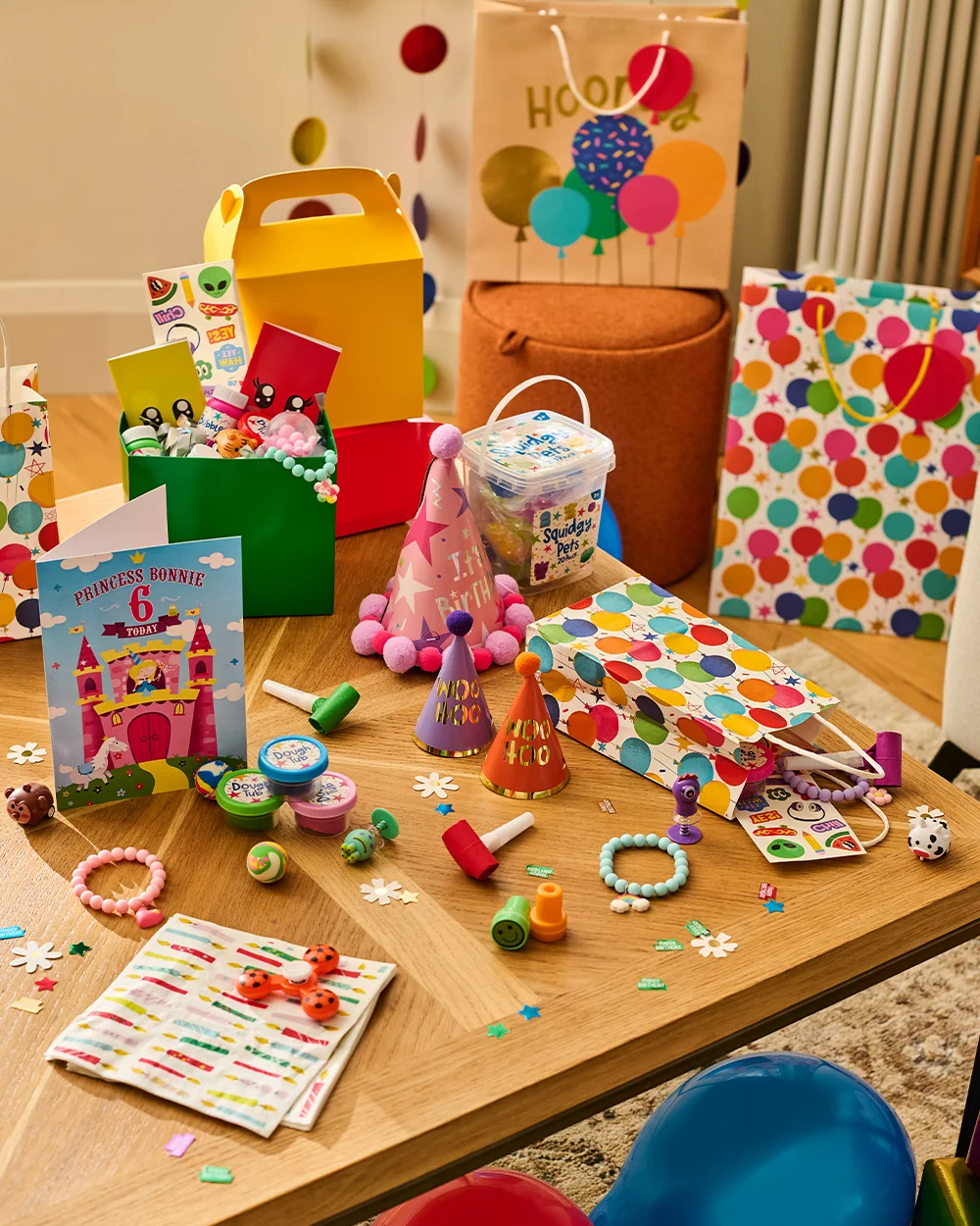 A children's party table covered with colourful party bag fillers and birthday accessories, including party hats, gift bags, favour boxes, sticker sheets, bracelets, modelling dough, slime tubs, novelty toys and sweet treats. A personalised birthday card reading ‘Princess Bonnie 6 Today’ stands among the items, with confetti and balloons scattered around the display.