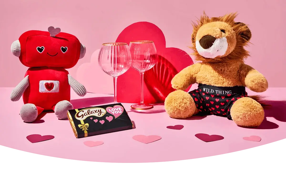 A cute Valentine’s display featuring a red robot plush, a lion plush with heart‑patterned shorts, two wine glasses, a chocolate bar, and a Valentine’s card arranged on a pink background with scattered paper hearts.