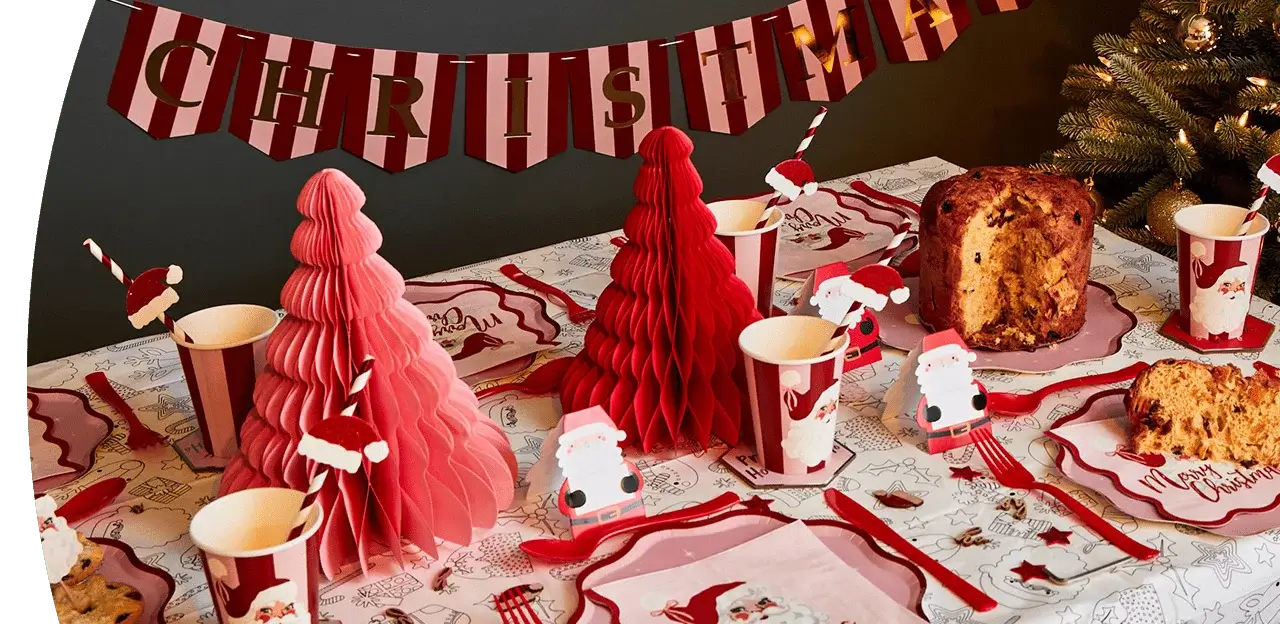 Christmas-themed party table with red and pink paper trees, Santa decorations, striped cups, festive plates, and a loaf of panettone under a red and white Christmas banner. Christmas-themed party table with red and pink paper trees, Santa decorations, striped cups, festive plates, and a loaf of panettone under a red and white Christmas banner.