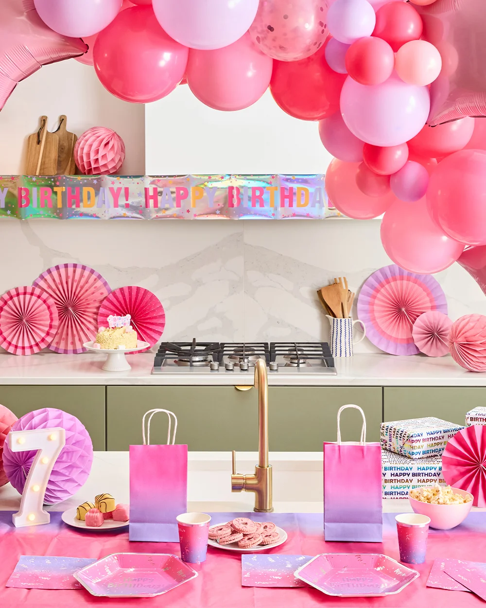 A pink-themed birthday party table set in a modern kitchen, decorated with a large pink balloon garland, star-shaped foil balloons, pink paper fan decorations and a holographic ‘Happy Birthday!’ banner. The table is laid with pink and lilac party plates, cups and napkins, alongside gift bags, wrapped presents, a bowl of popcorn, biscuits and a light-up number 7 decoration.