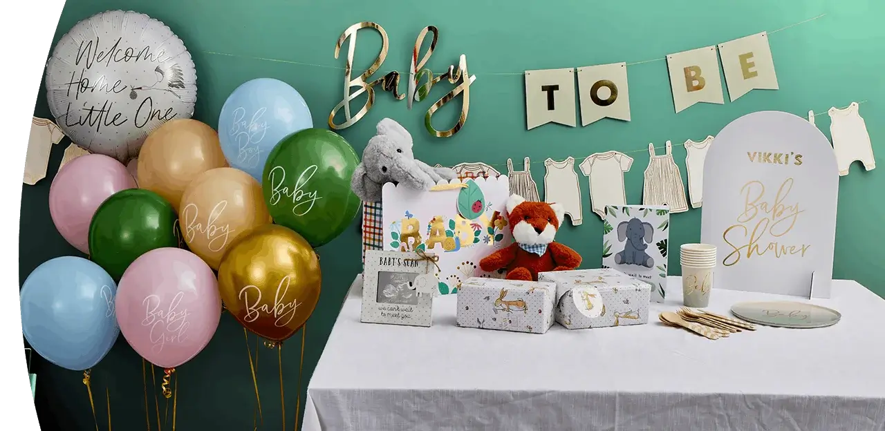 A baby shower table display with gift bags, plush toys, wrapped presents, cups, plates, and gold decorations, surrounded by colorful balloons and a banner reading Baby To Be.