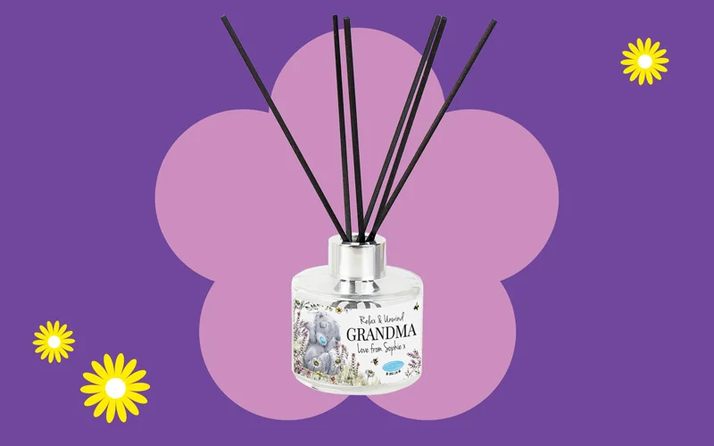 A floral‑themed reed diffuser with black reeds and a label reading ‘Relax & Unwind Grandma,’ displayed against a purple background with a pink flower shape and small yellow daisies.