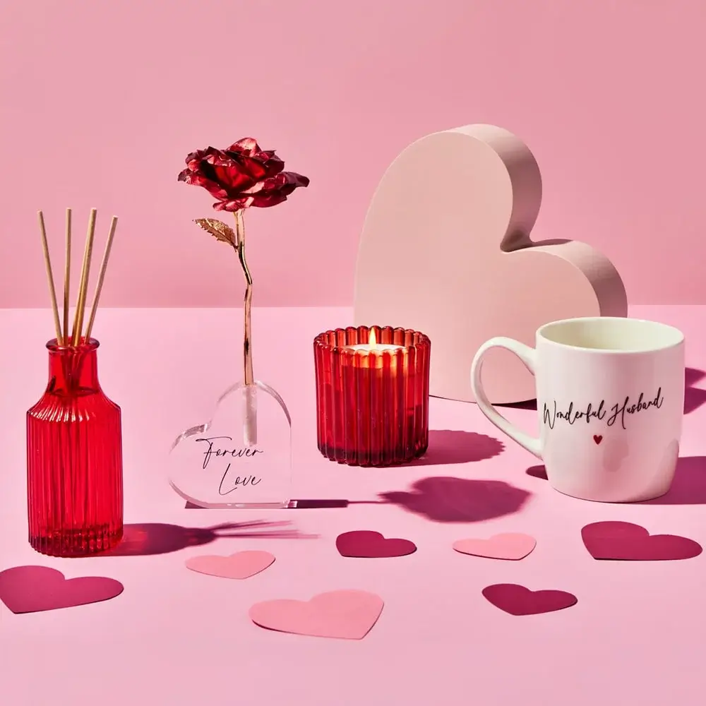 Valentine-themed arrangement featuring a red reed diffuser, a glass vase with a red rose labeled Forever Love, a red ribbed candle holder, a white mug with cursive text, and scattered paper hearts on a pink surface with a large heart-shaped decoration in the background.