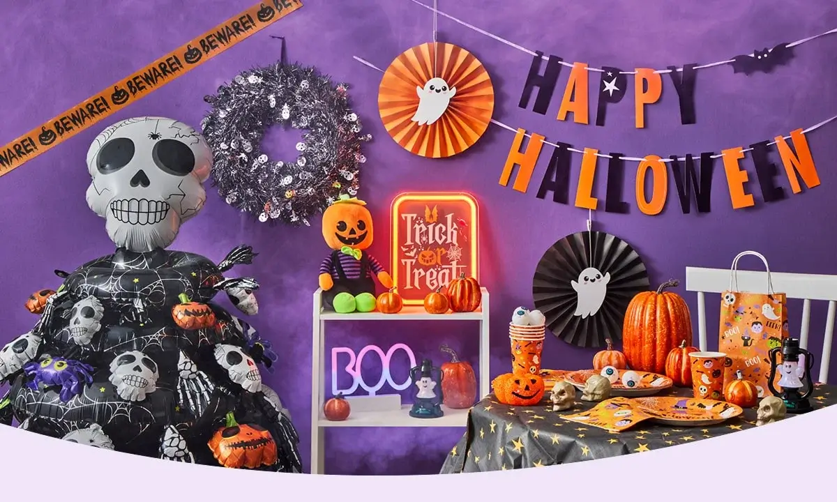 Halloween-themed party setup with a purple backdrop, black netting, paper bats, and garlands featuring pumpkins, skulls, and cat faces, centered around a large black cat face decoration with BOO and SPOOKY text, and a table set with themed plates, cups, candy, and spooky decorations.