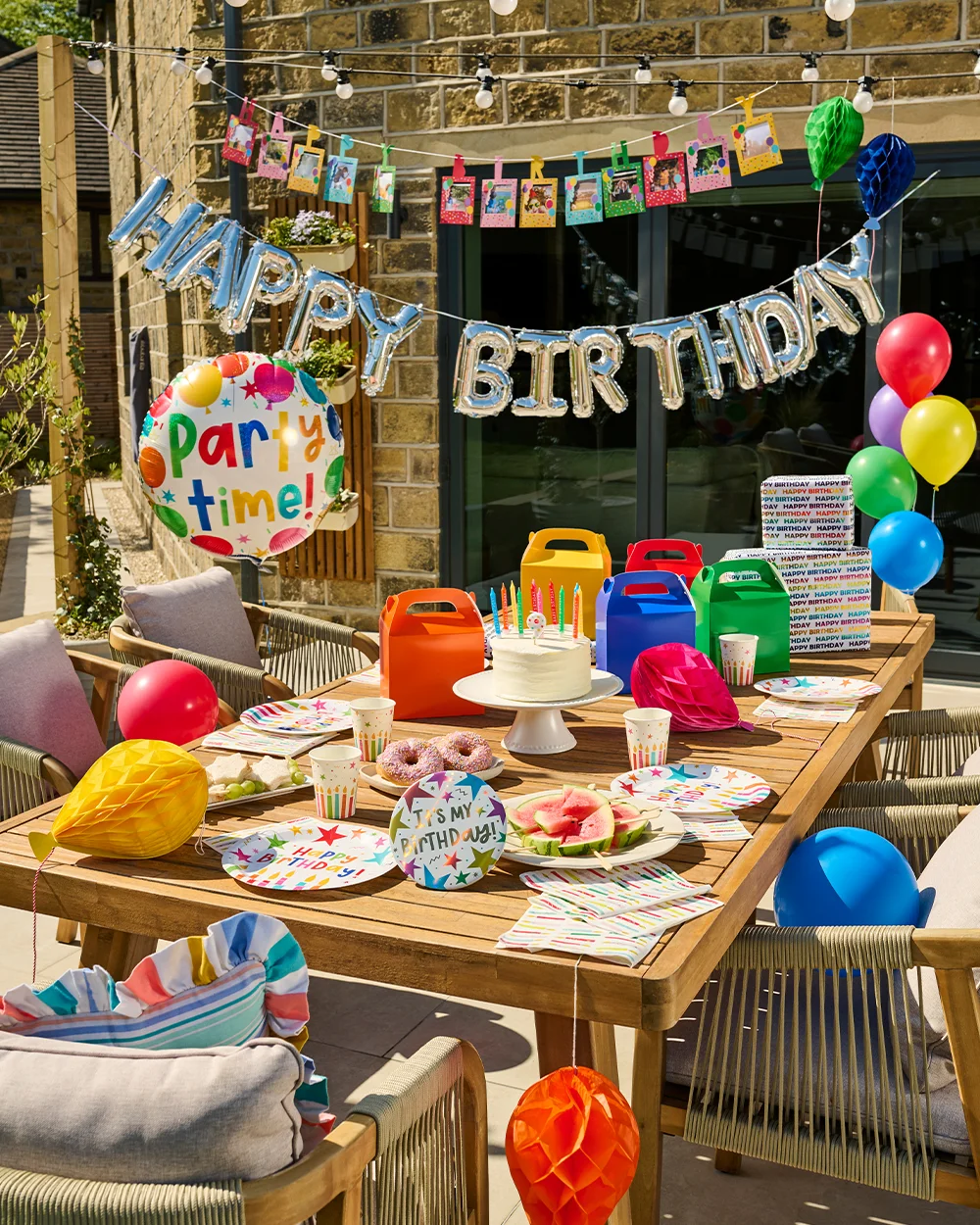 A colourful outdoor children's birthday party setup on a patio, featuring a wooden table decorated with rainbow-striped partyware, gift boxes, balloons and honeycomb decorations. A birthday cake topped with candles sits at the centre of the table, surrounded by doughnuts, fruit and party treats. Silver foil ‘Happy Birthday’ balloons hang above, alongside a photo bunting display, while brightly coloured balloons and a large ‘Party Time!’ balloon complete the festive garden celebration scene. A colourful outdoor children's birthday party setup on a patio, featuring a wooden table decorated with rainbow-striped partyware, gift boxes, balloons and honeycomb decorations. A birthday cake topped with candles sits at the centre of the table, surrounded by doughnuts, fruit and party treats. Silver foil ‘Happy Birthday’ balloons hang above, alongside a photo bunting display, while brightly coloured balloons and a large ‘Party Time!’ balloon complete the festive garden celebration scene.