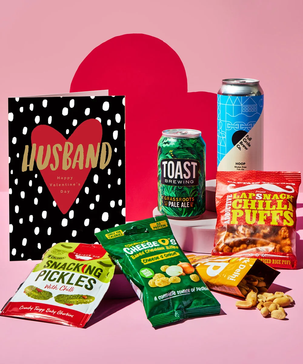 Valentine’s gift set displayed on a pink background with red heart-shaped props. The set includes a brown card with the text I ❤️ your and a rooster illustration, two cans of craft beer, and assorted snacks such as snacking pickles, cheese bites, chili puffs, and nuts in colorful packaging.