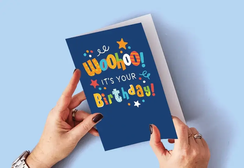 A person holding a blue birthday card with colorful text that says Woohoo! It's your Birthday, decorated with stars, dots, and squiggles.