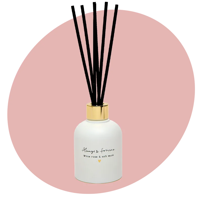 A white reed diffuser bottle with a gold lid and black reeds, labelled ‘Always & forever – white rose & soft musk,’ displayed against a soft pink circular background. A white reed diffuser bottle with a gold lid and black reeds, labelled ‘Always & forever – white rose & soft musk,’ displayed against a soft pink circular background.
